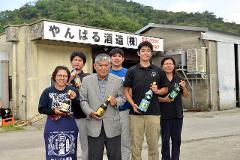 6명의 사람들이 &quot;얀바루 주조(주식회사)&quot;라고 적힌 간판이 있는 건물 앞에 서 있는데, 그들은 오키나와의 지역 술인 아와모리가 담긴 병을 들고 있습니다.