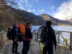Four men wearing backpacks and hiking gear stand on a wooden dock overlooking a serene lake and tree-covered mountains under a partly cloudy sky.