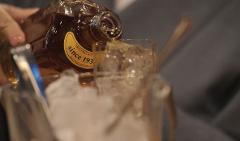 A hand pours amber liquid from a bottle labeled "Since 1936" into a glass filled with ice cubes.