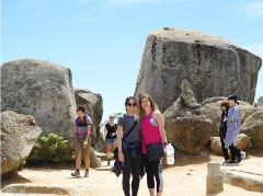 Two young women pose in front of large rocks with other people walking around them on a sunny day.