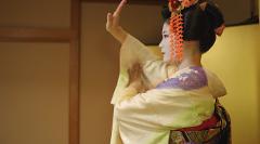 A geisha in a traditional kimono and elaborate hair ornaments performs a graceful dance in a room with gold and wood-paneled walls.