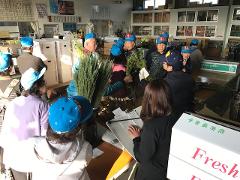A group of people wearing blue hats are gathered indoors, possibly at a flower market, with some holding bunches of flowers and others taking notes.