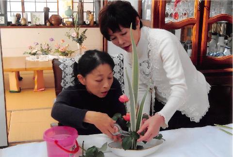 Two women are arranging flowers. One is seated and cutting a rose stem with clippers, while the other stands behind, guiding the placement of a tall green leaf.