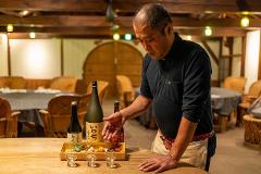 A man in a black polo shirt and apron presents a tasting set of sake bottles and small food pairings on a wooden table in a restaurant.