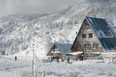 In a serene winter landscape, a person in an orange jacket walks along a snowy path towards a rustic wooden cabin nestled at the foot of a snow-covered mountain.