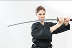 A woman in a black gi holds a katana sword horizontally in front of her on a white background.