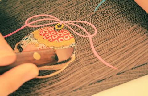 A close-up shows a hand holding a wooden tool over a decorative object with fabric and string elements on a textured surface.
