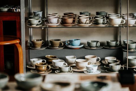 Pottery & Porcelain: Stories of the Artisans Behind the Craft A collection of ceramic cups and saucers in various earthy tones and styles are displayed on shelves in a glass cabinet.