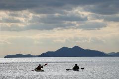 Two kayakers paddle on a shimmering body of water towards distant mountains under a cloudy sky.