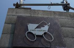 A sign reading "ONBA FACTORY" with a stroller icon is mounted on a brick wall, with scaffolding visible above against a blue sky.