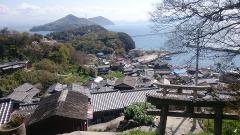 A scenic view overlooking a traditional Japanese village with tiled roofs, leading to a harbor and the sea, framed by mountains in the distance and a torii gate in the foreground.