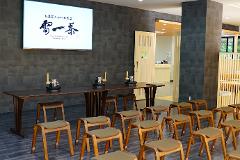 This image shows a modern tea room with a large screen displaying Japanese calligraphy, long tables, and rows of wooden stools, suggesting a space for tea ceremonies.