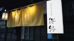 The image features a Japanese restaurant called "Sakura-michi", adorned with traditional yellow noren curtains and a vertical sign with Japanese calligraphy.