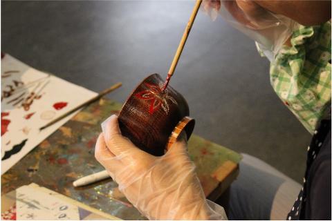 A gloved hand delicately paints a red flower onto a lacquered bowl with a thin brush.
