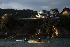 Two kayakers paddle in the calm waters near a rocky shoreline with trees, a traditional Japanese building, and a modern house under a cloudy sky.