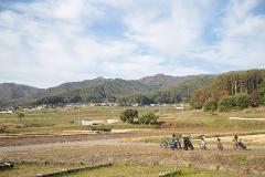 A serene landscape of rolling hills and farmland under a bright, cloudy sky, dotted with a small village and a line of parked bicycles.