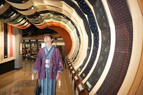 Nagano Cultural Day Tour: Ina Tsumugi Kimono & Textile Craft A person dressed in traditional Japanese attire gazes up at a tunnel-like display of intricately woven silk fabrics in a museum.