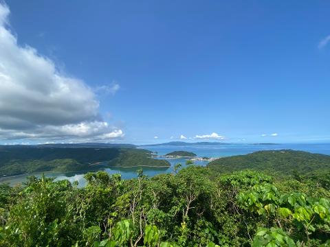 A scenic vista of Shioya Bay, Okinawa, with lush green islands and a turquoise sea under a partly cloudy blue sky, showcasing the natural beauty of the "Village of Longevity".