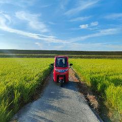 A red electric tuk-tuk is parked on a rural road surrounded by vibrant green rice fields under a clear blue sky with wispy clouds.