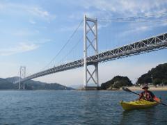 A person in a yellow kayak paddles on a calm body of water with a large white suspension bridge in the background.