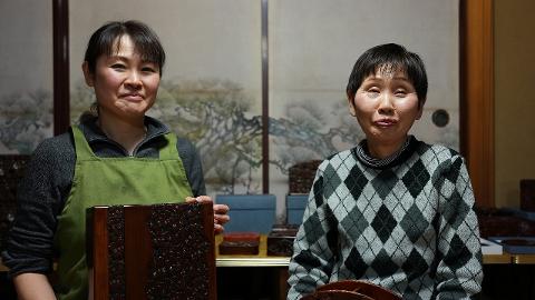 Two women in traditional Japanese attire proudly display intricately carved wooden art.