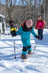 A child in a turquoise snowsuit and pink hat snowshoes through a snowy forest, holding ski poles, with other people and a van in the background.