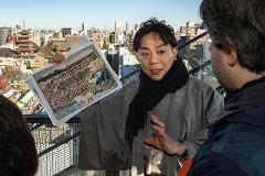 A man in a grey robe and black scarf holds up a historic map of Asakusa to another man in a blue jacket, with the city skyline in the background.