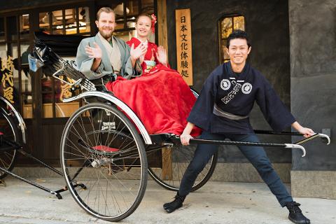 A smiling man pulls a rickshaw with a couple dressed in traditional Japanese clothing sitting inside, in front of a building with Japanese writing on it.
