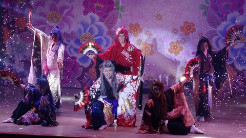A group of six performers in traditional Japanese clothing and colorful wigs pose on stage during a performance with fans in hand, confetti falling around them.