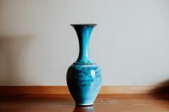 Pottery & Porcelain: Stories of the Artisans Behind the Craft A tall, slender turquoise vase with a glossy finish sits on a wooden surface against a light grey wall.