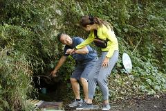 A man and a woman are laughing while looking at water trickling down a mossy rock face in a forest.