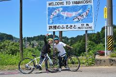 Two cyclists in helmets take a selfie with bicycles in front of a sign that reads "Welcome to Yamakoshi" with koi fish imagery.