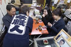 Four people are gathered around an orange table, engaged in the process of crafting hand-carved seals, with two women in the foreground holding tools and appearing to work on the seals.