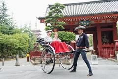 A smiling rickshaw driver pulls a rickshaw with two tourists in front of a temple in Asakusa, Tokyo.