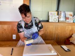 A person wearing blue gloves and a plastic apron cuts a block of white material with a knife on a white cutting board.