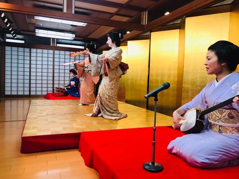 A cultural performance featuring geishas dancing with fans and a musician playing a traditional instrument on a red platform.
