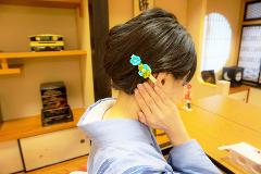 A woman in a blue kimono styles her hair with a tsumami-zaiku accessory in a traditional Japanese setting.