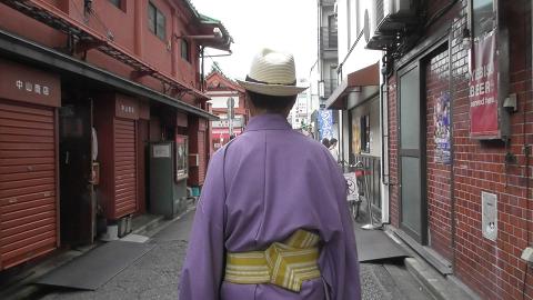 A person in a purple kimono and straw hat walks down a narrow street lined with traditional Japanese buildings and shops.