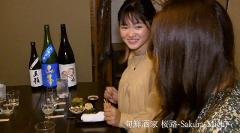 Two women are seated at a table with sake bottles, tasting glasses, and appetizers at what looks like a bar in Hakodate.
