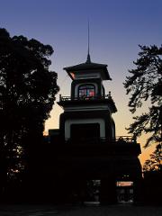 A dark silhouette of a bell tower with stained glass windows stands against a twilight sky, framed by trees.