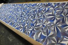 A long textile with a repeating abstract floral pattern in shades of blue and brown is stretched across a wooden frame.