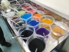 Clear plastic bowls filled with various colorful liquids and spoons are displayed on a stainless steel surface.