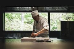 A chef in a white uniform prepares food on a cutting board with a large window overlooking lush greenery in the background.