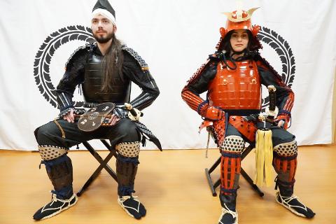 A man and a woman dressed in traditional samurai armor sit on chairs in front of a white backdrop with a black circular emblem.
