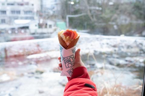 A hand in a red jacket holds a Tote-yaki crepe with strawberries and cream, with a snowy outdoor scene visible through a window.