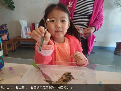 A young girl proudly displays a pearl she extracted from an oyster, with a pink jacket and a tray containing the oyster remains in front of her.