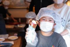 A young boy wearing a hairnet and face mask holds a crepe with strawberries and whipped cream.