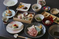 A traditional Japanese meal featuring an assortment of small dishes, including grilled meat, soba noodles, rice, and various appetizers, is artfully arranged on a dark surface.