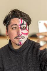 A person with half of their face painted in Kabuki style holds up a smartphone to take a selfie.