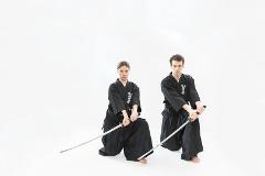 Two people in traditional samurai attire kneel with swords drawn against a white background.
